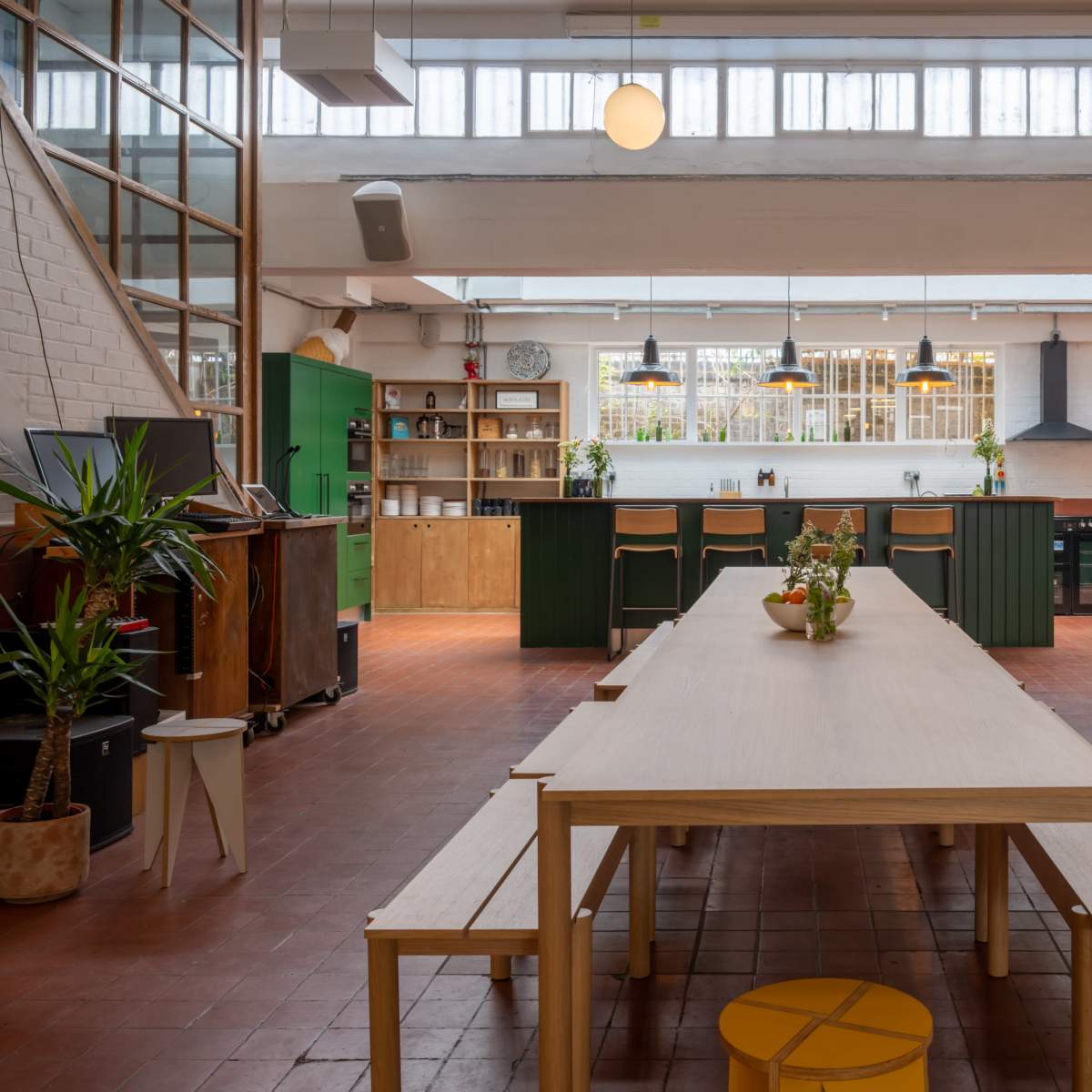 A spacious kitchen and dining area with a long wooden table, benches, green cabinetry, and large windows allowing natural light to fill the space.