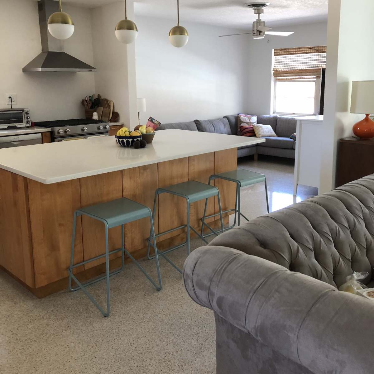 A modern kitchen with a central island and bar stools, connected to a living area furnished with a gray sofa.