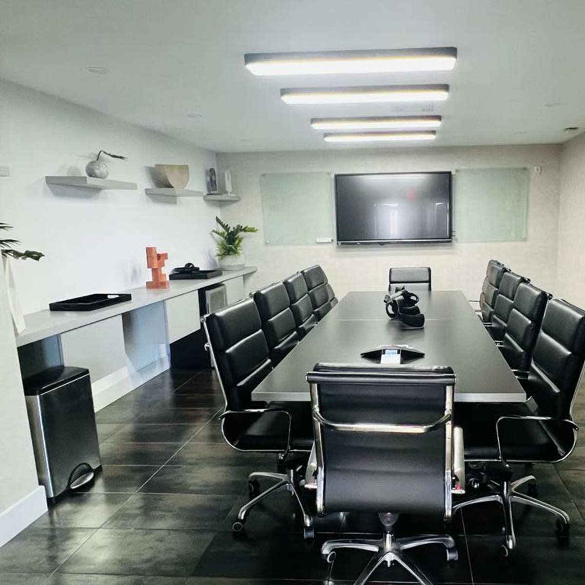 A conference room features a large rectangular table surrounded by black leather chairs, with a television mounted on the wall and a painting beside it.