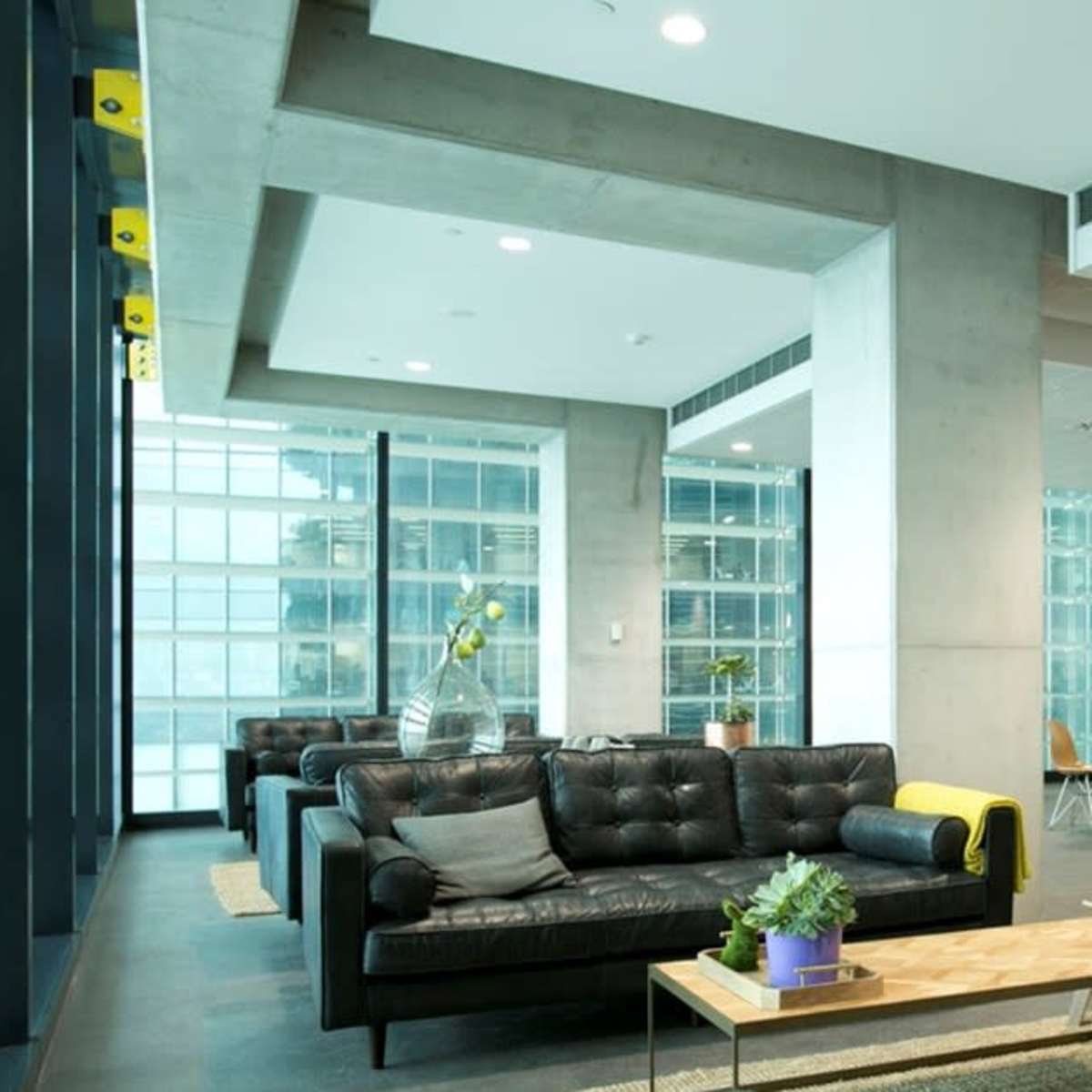 A modern office lounge with black leather sofas, a wooden coffee table, and large windows providing views of a glass building.