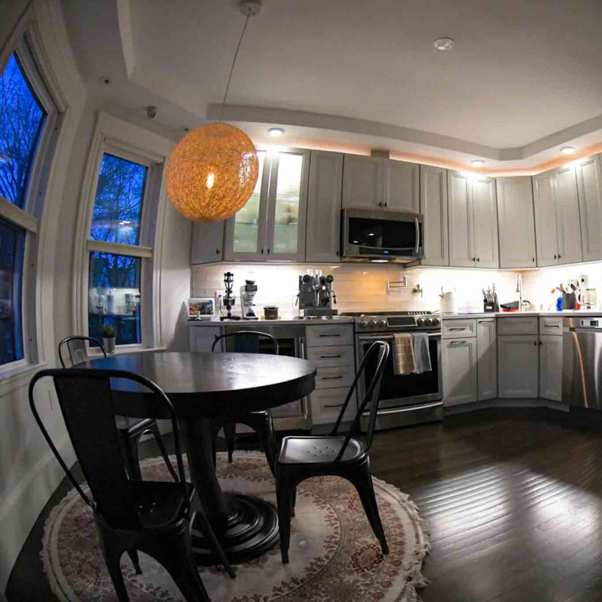 A modern kitchen features stainless steel appliances, white cabinetry, a round table with black chairs, and a large pendant light.