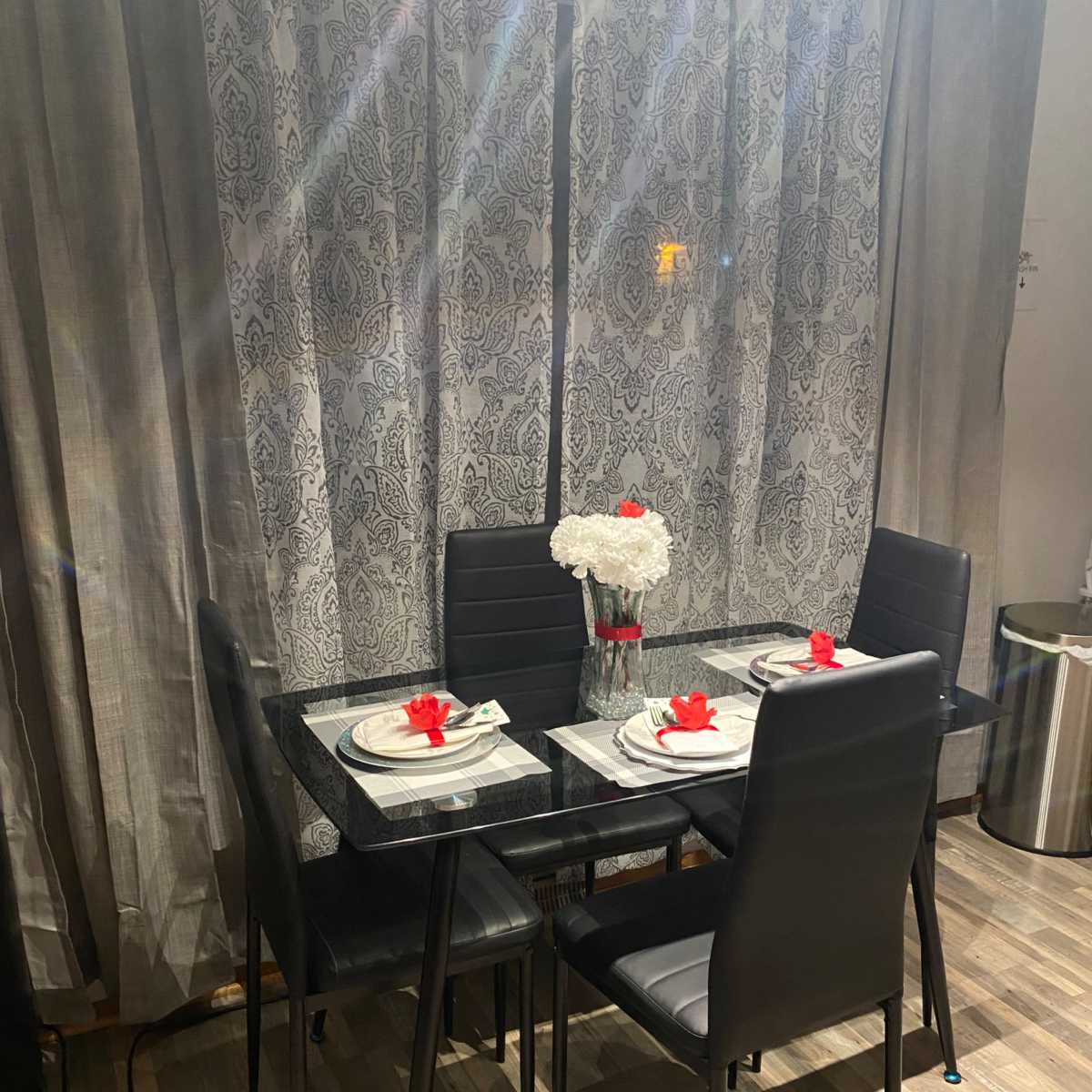 A glass-topped dining table is set with plates and red flowers in a vase, surrounded by four black chairs in front of patterned curtains.