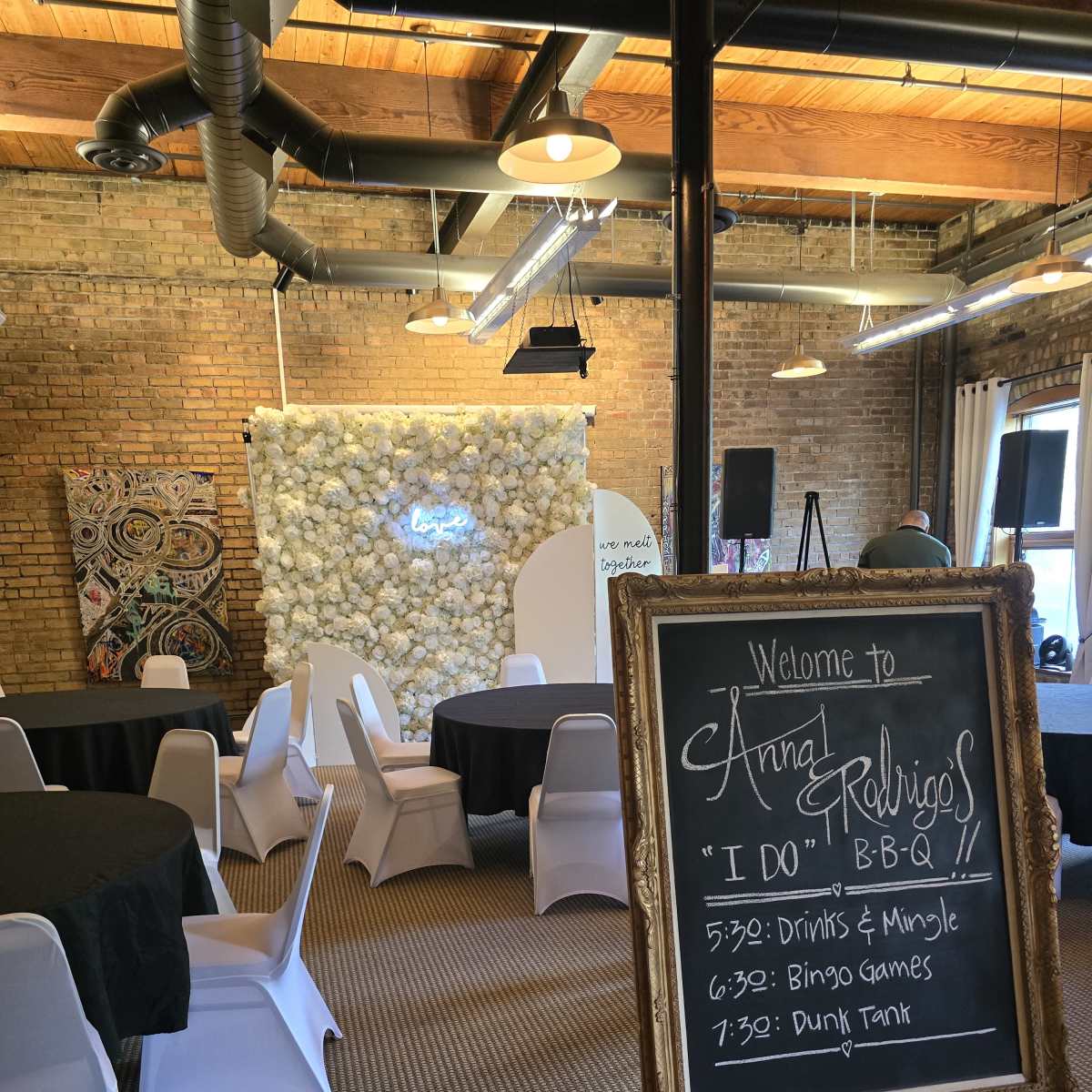 An event space with round tables covered in black cloths, white chairs, and a decorative wall made of flowers, alongside a welcome sign detailing the schedule for a BBQ event.