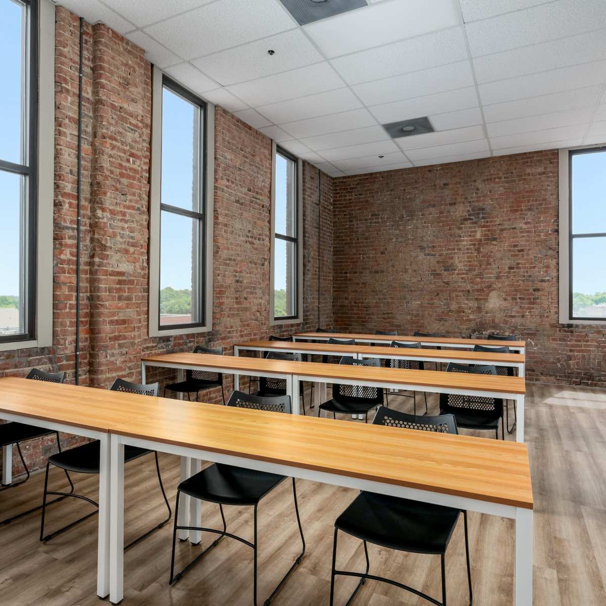 A classroom with multiple long tables and chairs set against a backdrop of exposed brick walls and large windows.