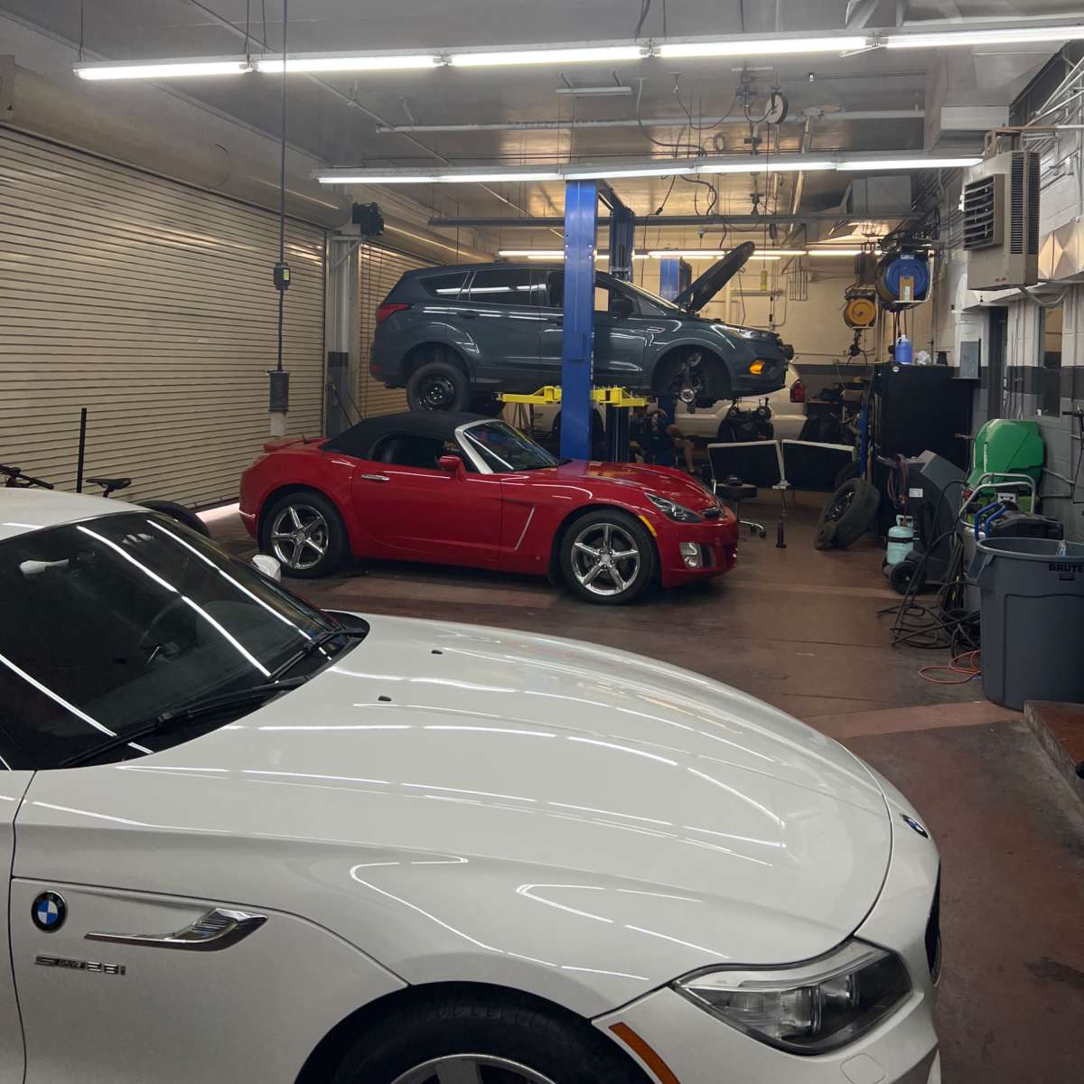 A car repair shop with a red convertible in the foreground, a white car on the left, and a blue vehicle being serviced on a lift in the background.
