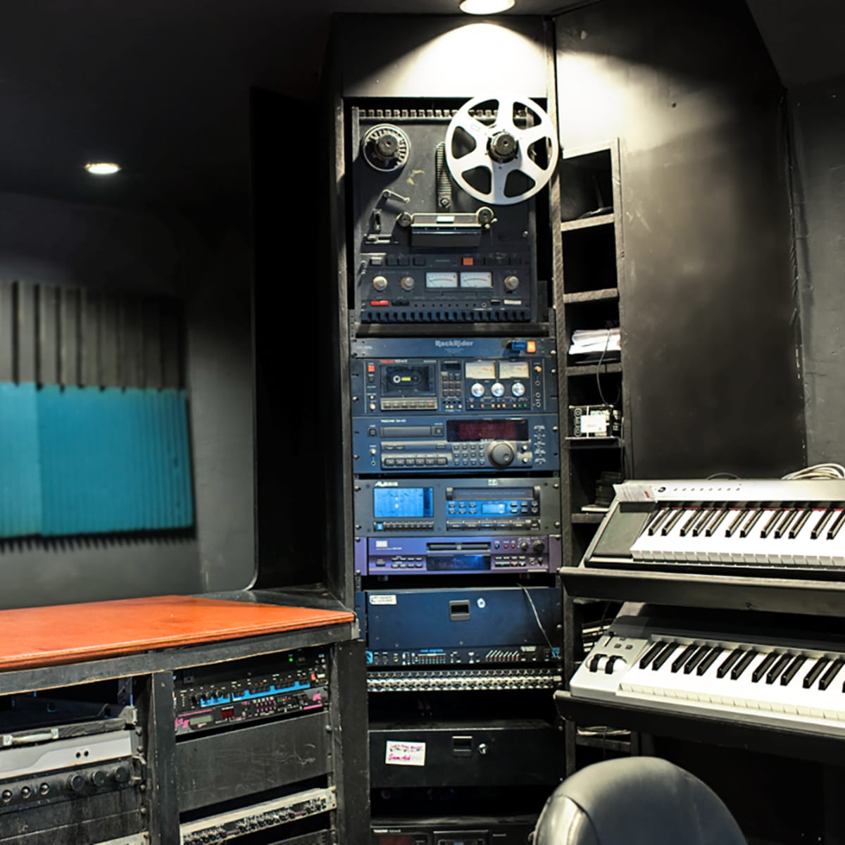 A music studio with a wooden tabletop, a vertical array of audio equipment, and a keyboard positioned next to a soundproof wall.