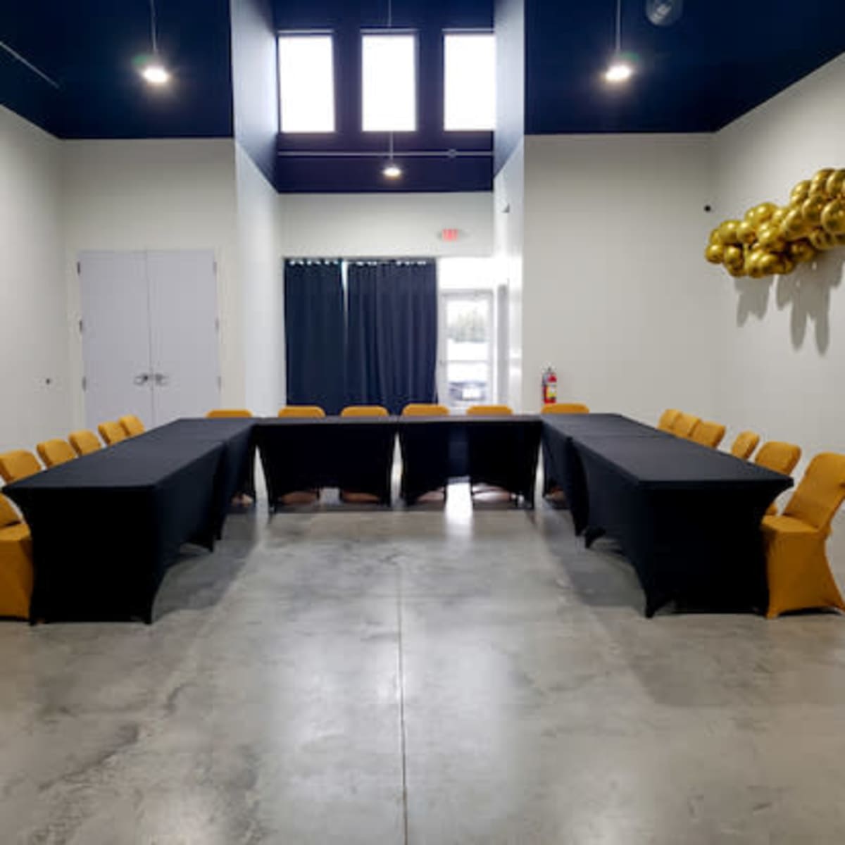 A conference room features two long tables arranged in an L-shape, surrounded by yellow chairs, with large windows and a wall decorated with gold balloons.