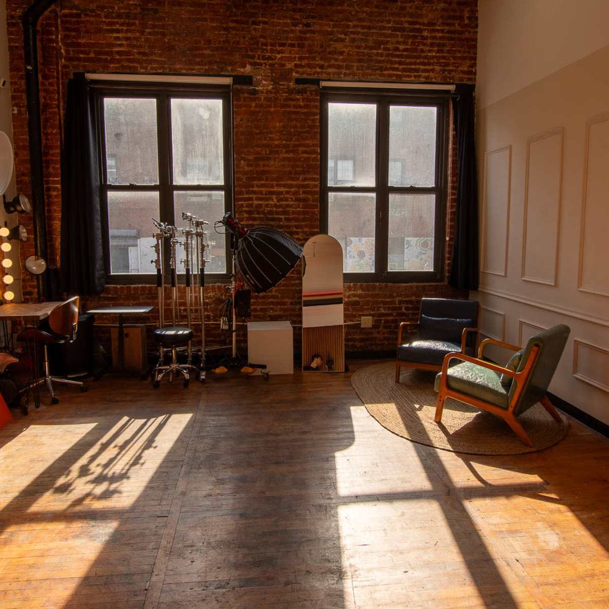 An interior studio space with exposed brick walls, large windows, a wooden floor, and various props including a chair, a desk with a mirror, and backdrop panels.