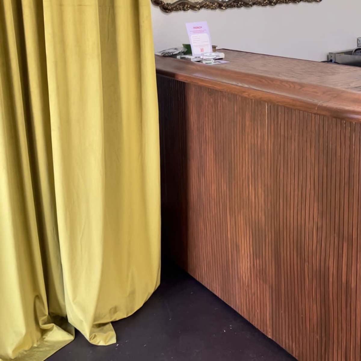 A wooden reception desk next to a long yellow curtain and a decorative mirror on the wall, with shelves displaying items above the desk.