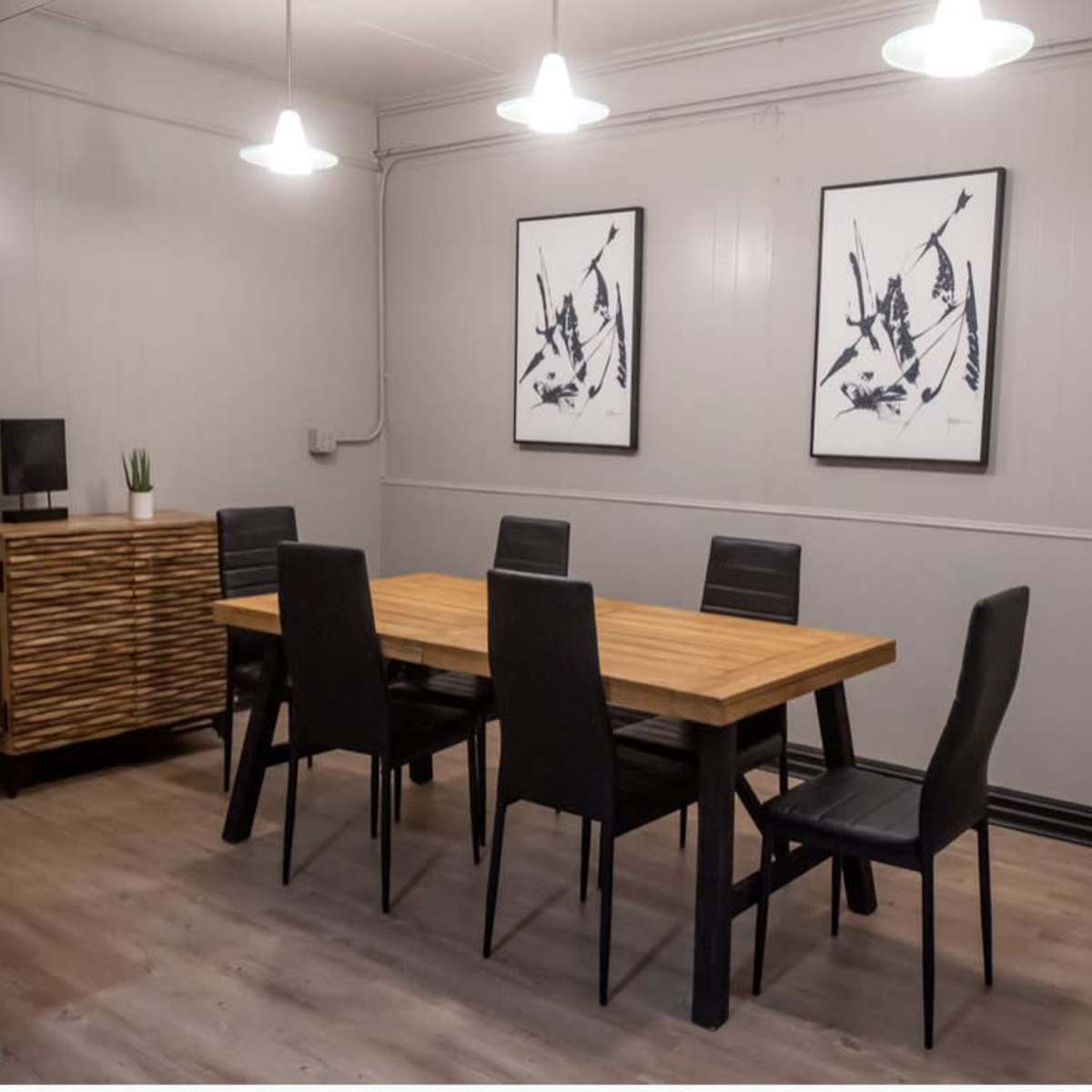 A dining area features a rectangular wooden table surrounded by six black chairs, with two framed artworks on the wall and overhead lighting.
