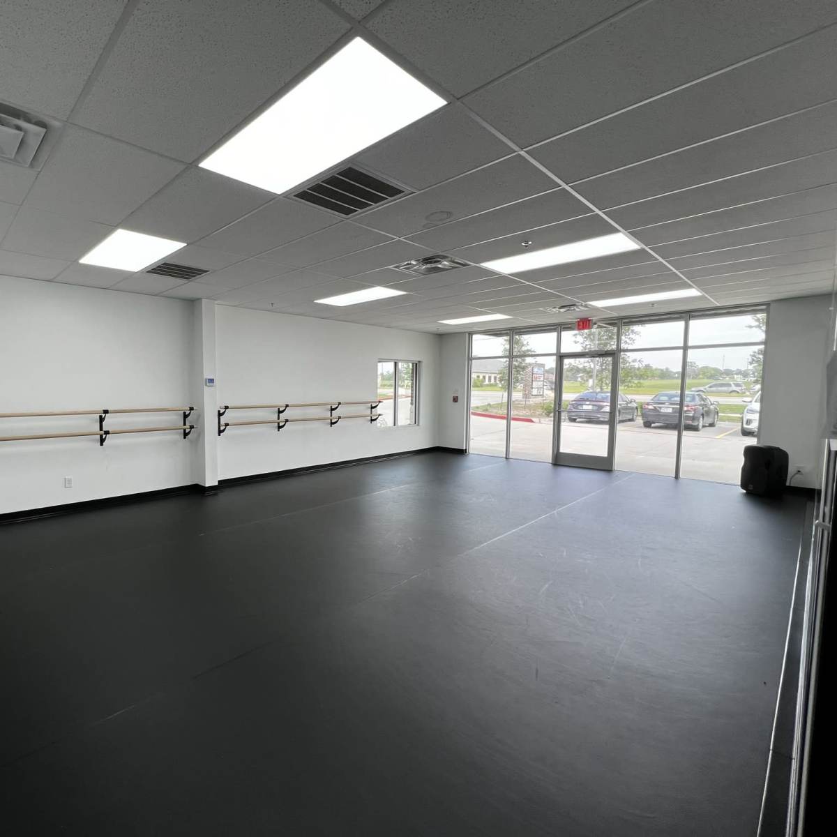 A spacious dance studio with a black floor, mirrored walls, and ballet barres along one side, along with large windows letting in natural light.