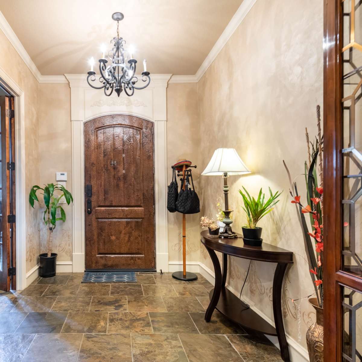 The entrance hall features a wooden door, a chandelier overhead, a table with plants, and a coat rack against a textured wall.