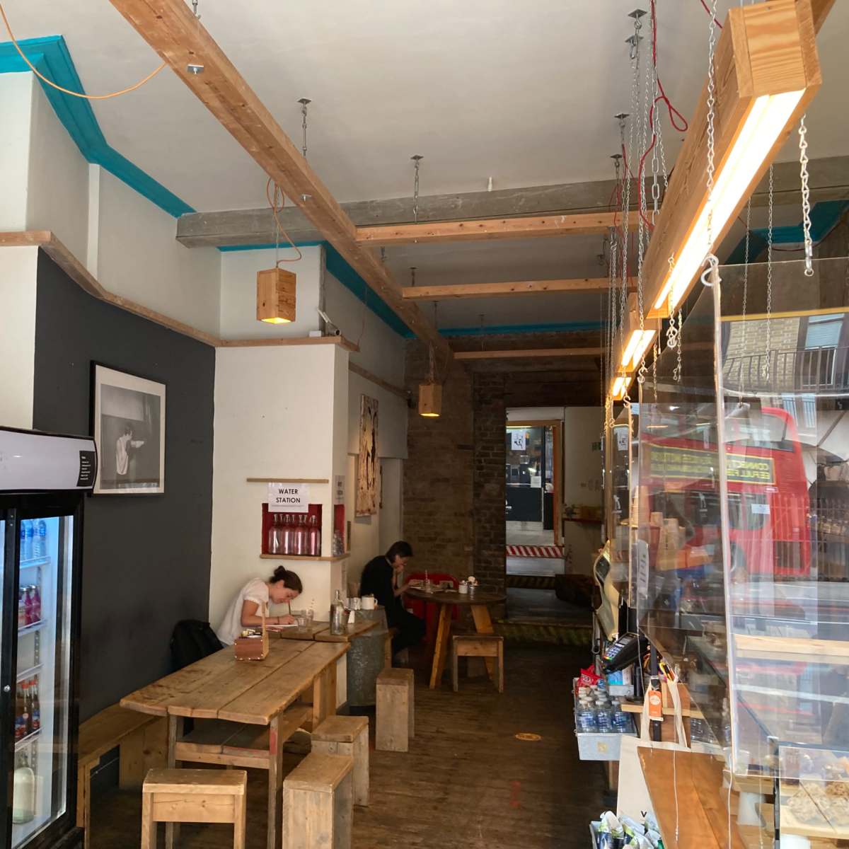 The interior of a café features wooden furniture, a glass display case, and a refrigerated drink cooler, with a person sitting at a table.