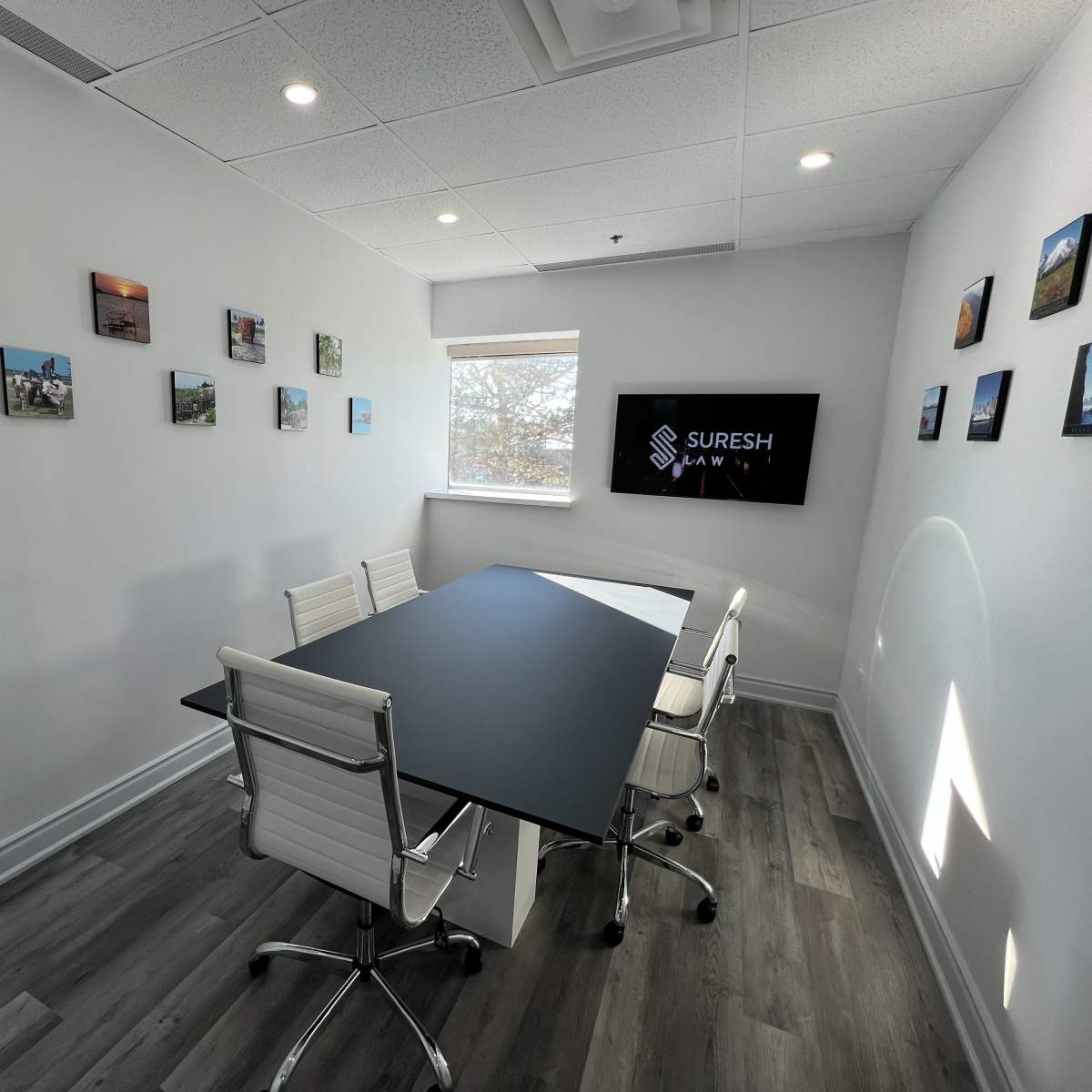 A modern conference room with a large black table, white office chairs, and framed photos hanging on the walls, along with a screen displaying the name "Suresh."