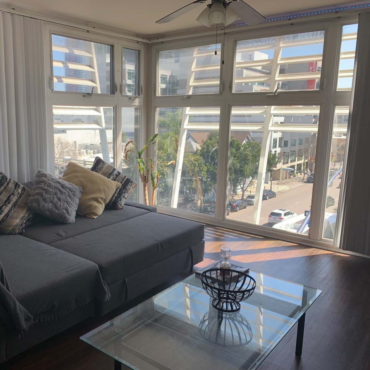 A gray couch with decorative pillows sits in a bright room with large windows overlooking a city street.