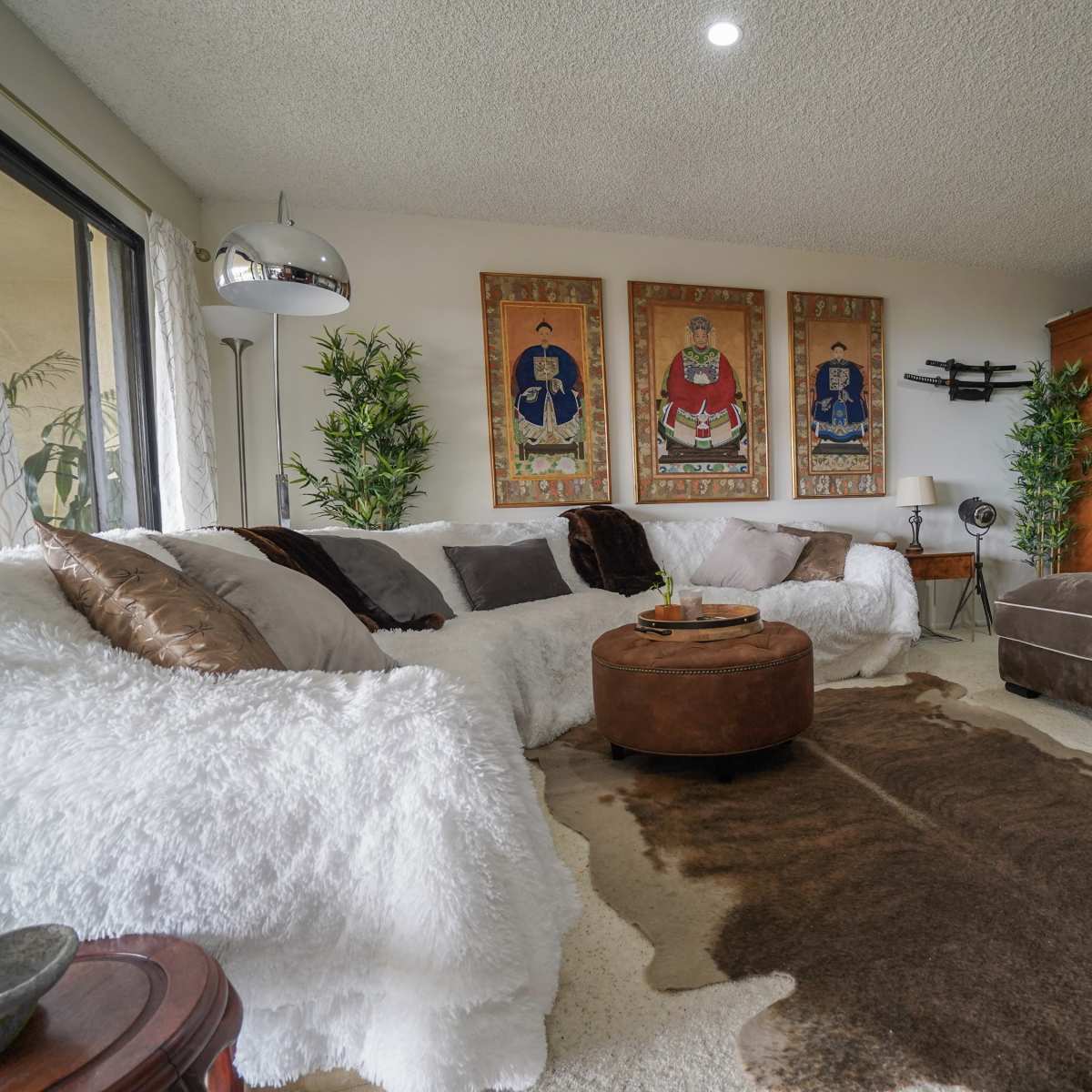 The living room features a sectional sofa, a round ottoman, and decorative wall art flanked by potted plants.