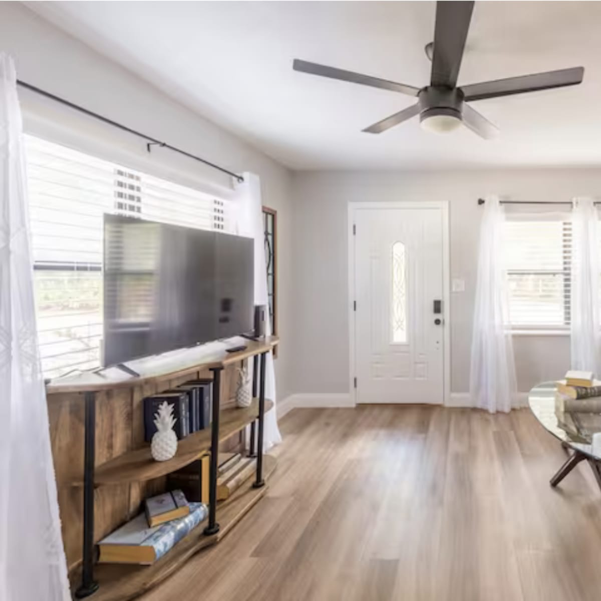 The living room features a dark leather sofa, a glass coffee table, a television on a wooden stand, and large windows with sheer white curtains.