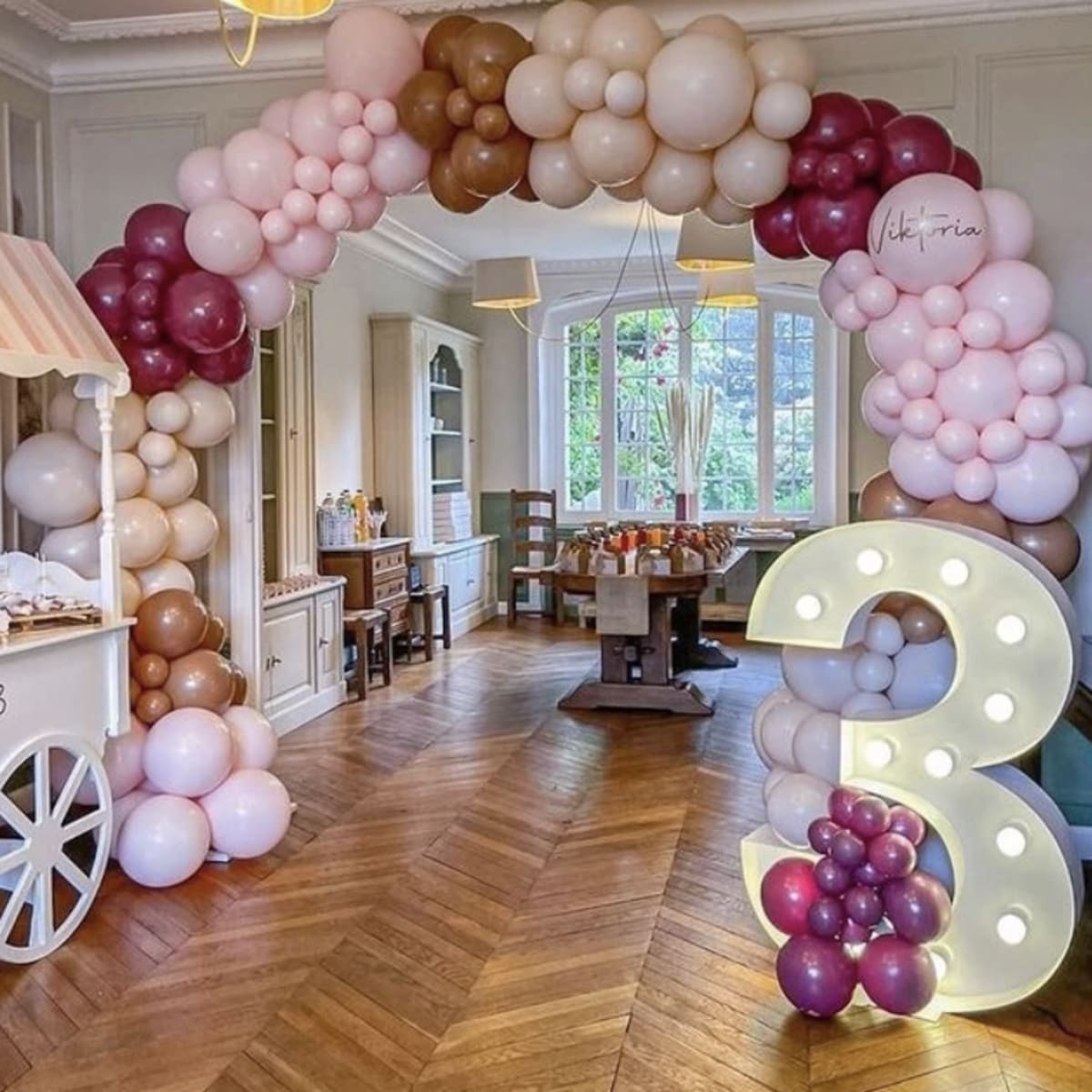 A decorative balloon arch in shades of pink, brown, and burgundy frames a large illuminated number three in a well-lit room with wooden flooring and furniture.