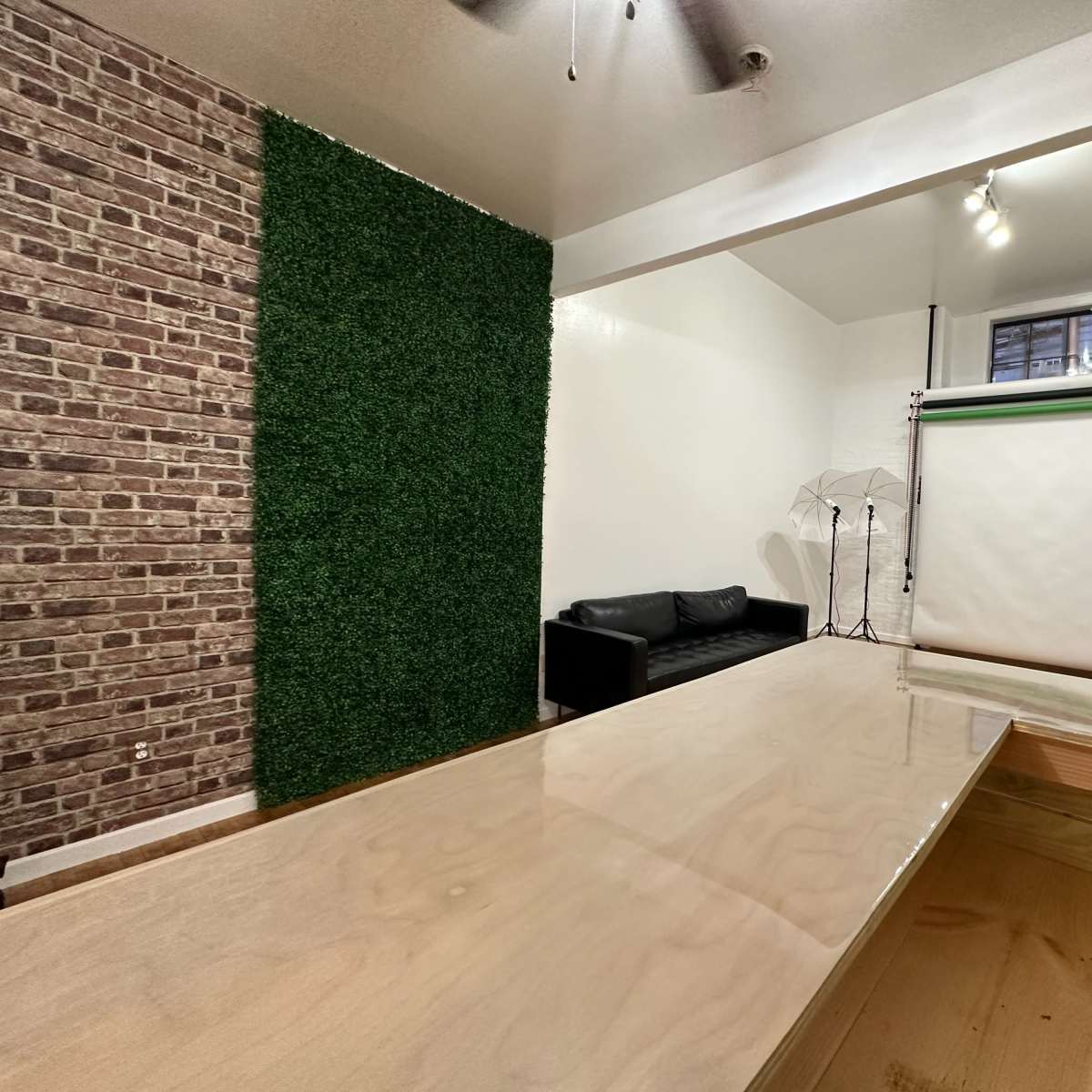 A modern studio space features a wooden table, a green wall, a black sofa, and photography equipment set up against a white backdrop.