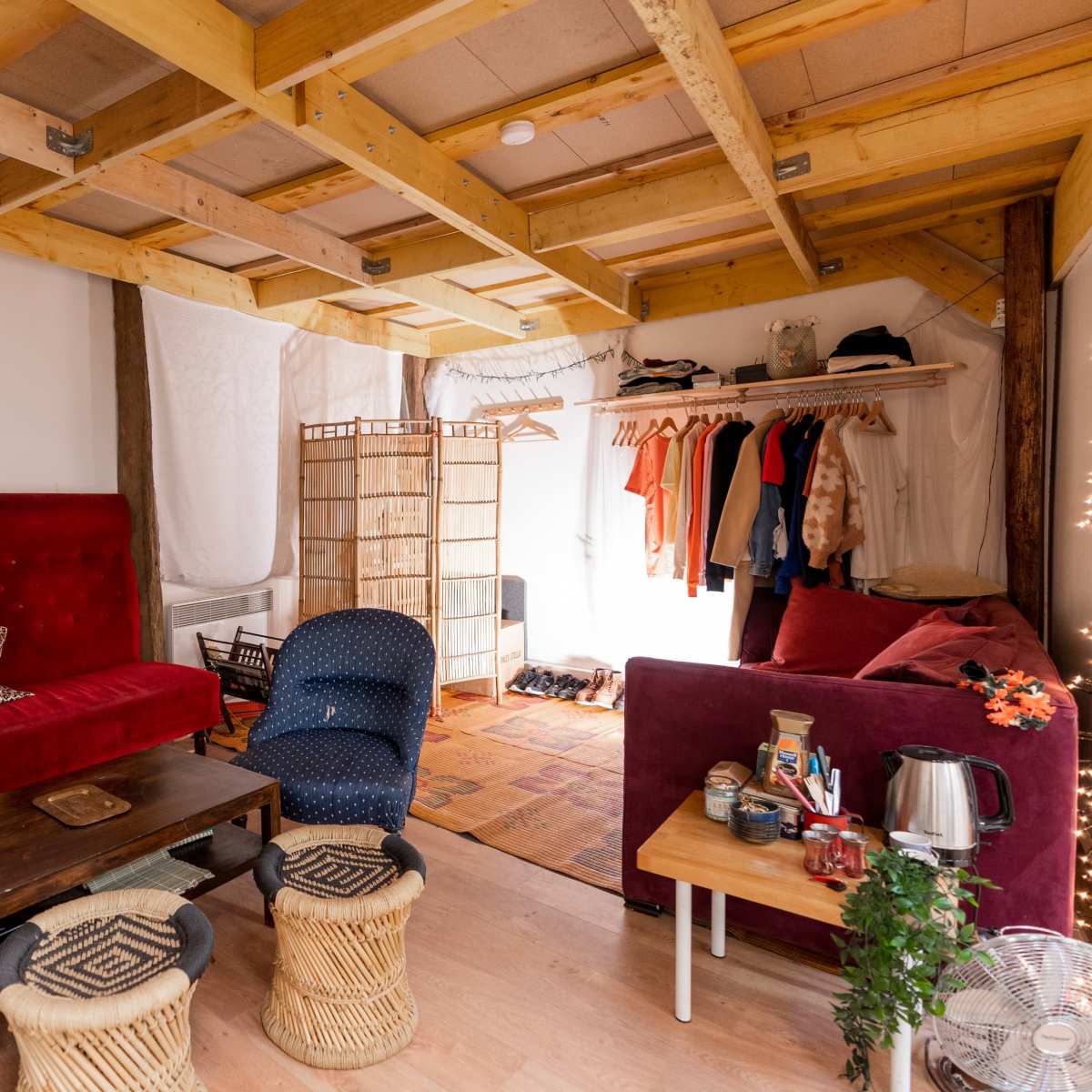 A cozy interior space with a red velvet sofa, a wooden table, and a clothing rack, complemented by natural wood beams and light-colored walls.