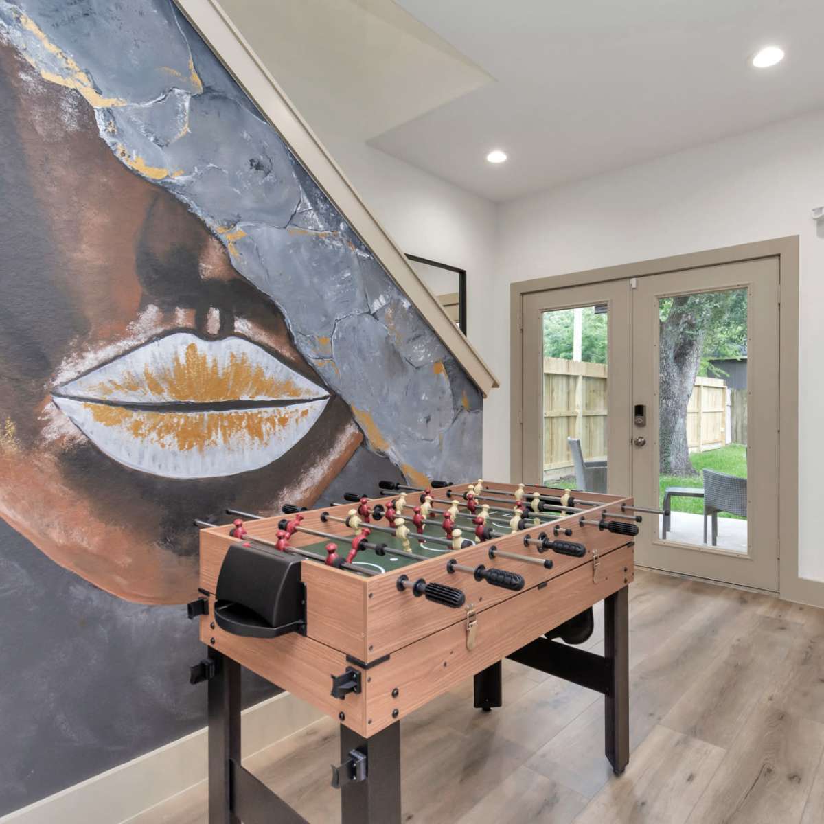 A foosball table is positioned near a staircase against a wall featuring a large mural of a face with striking gold accents.