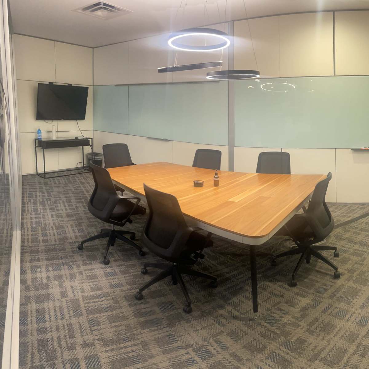 A modern conference room features a large wooden table surrounded by six ergonomic chairs, with a television and glass walls.