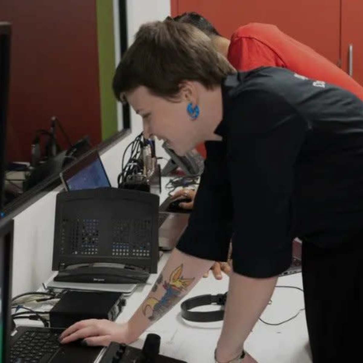 A person with short hair and visible tattoos leans over a desk filled with computer monitors and equipment while another individual works in the background.
