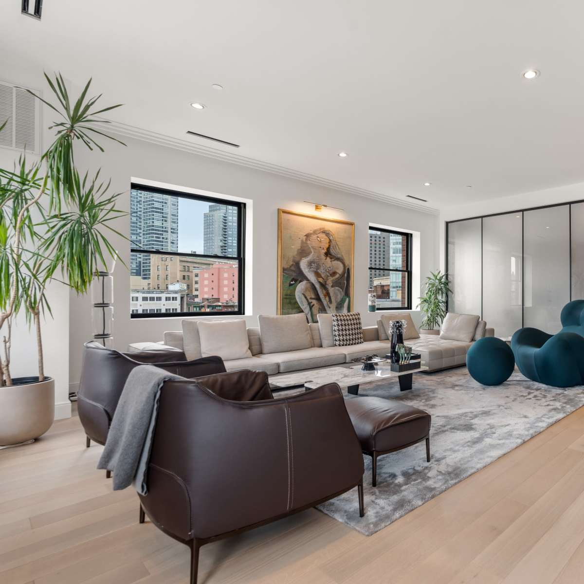 A modern living room with a large sectional sofa, a rug, and a glass-walled room in the background, featuring potted plants and artwork on the walls.
