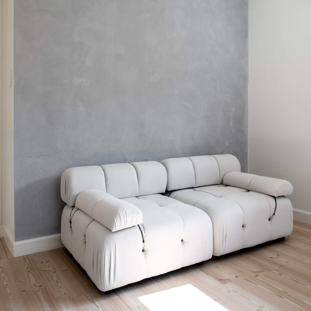 A white sectional sofa is positioned against a light gray textured wall in a minimalistic room with wooden flooring.