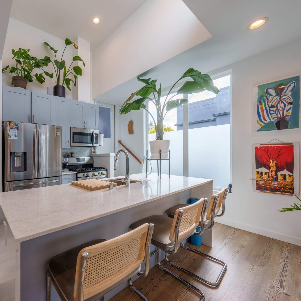 A modern kitchen features a large island with bar stools, stainless steel appliances, and abundant plants on the countertop and shelves.