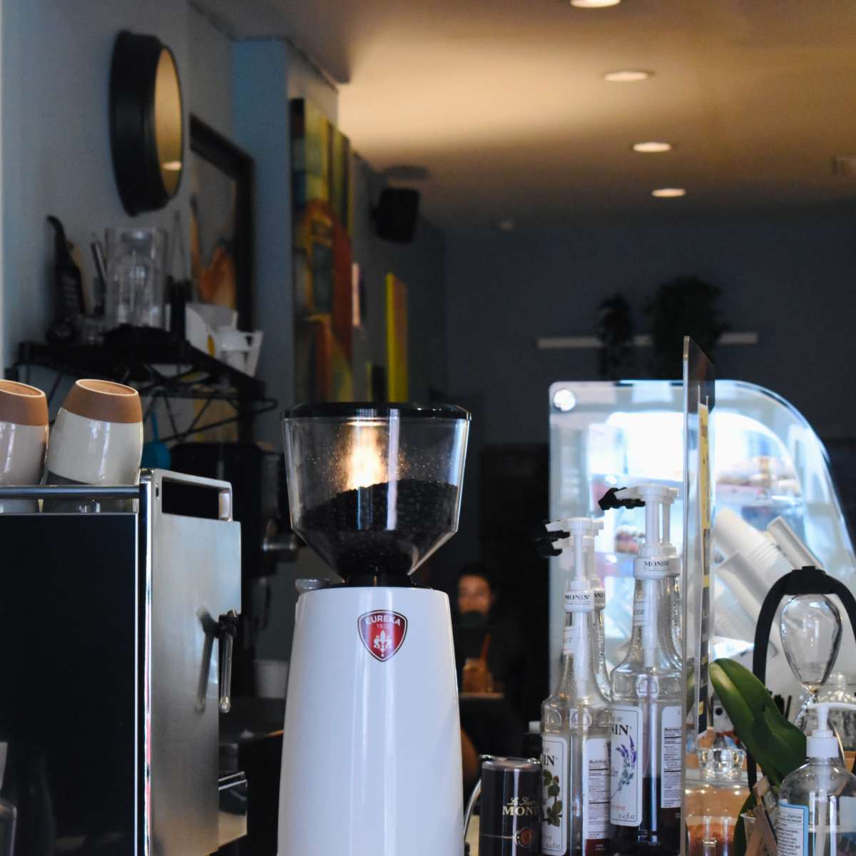 A coffee shop interior with an espresso machine, a coffee grinder, and neatly stacked cups on a counter, surrounded by shelves displaying menu items and pastries.