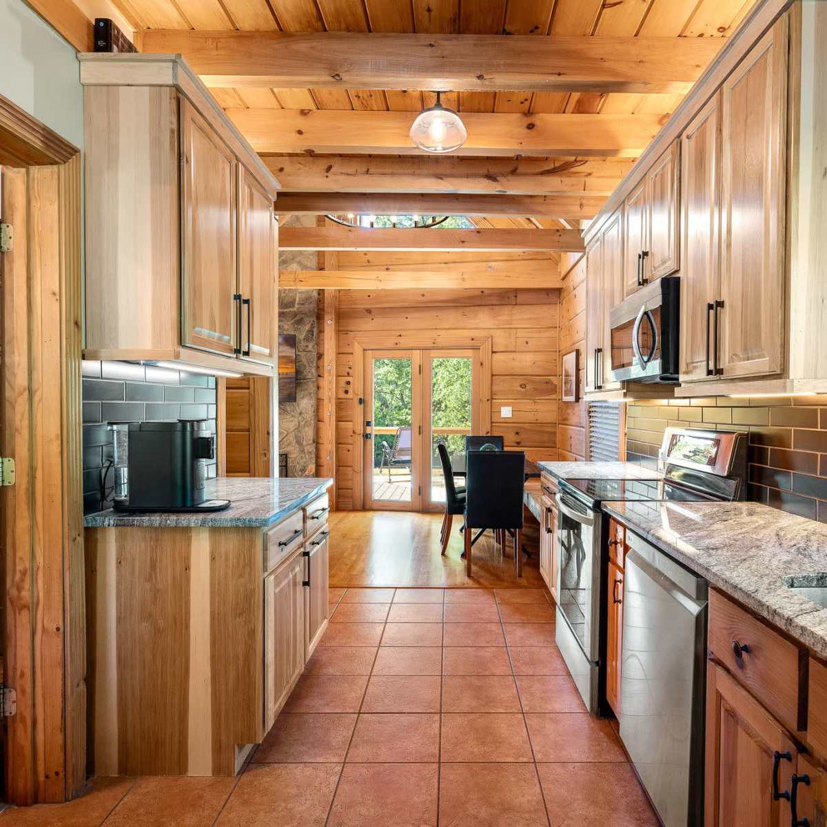 A rustic kitchen with wooden cabinets, granite countertops, and a view of a patio through a large window.