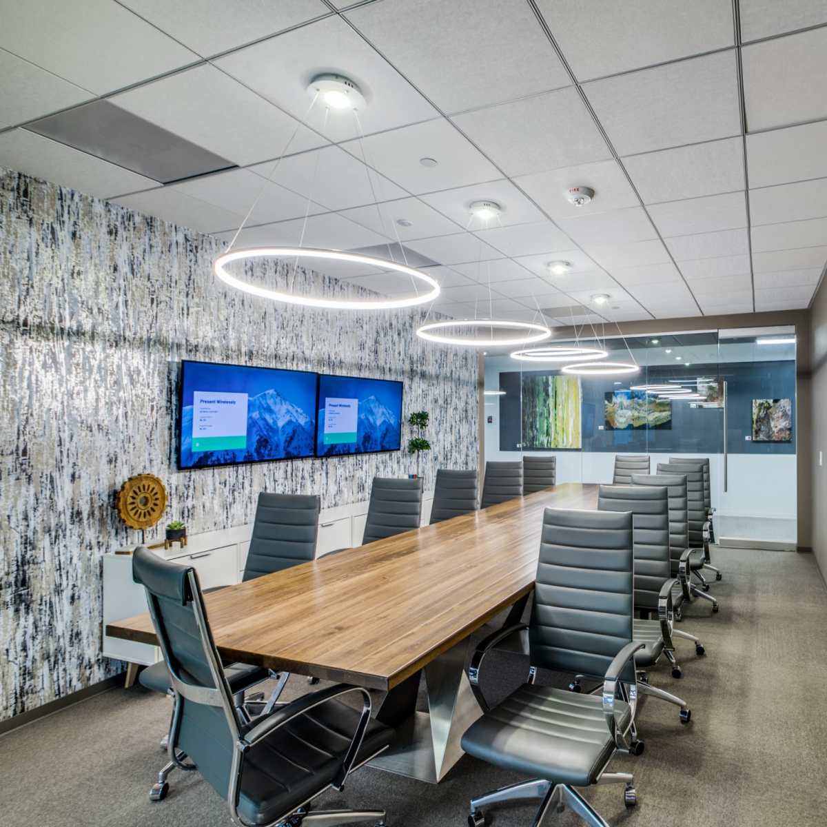 A modern conference room features a long wooden table surrounded by ergonomic chairs, with two wall-mounted screens displaying presentations.