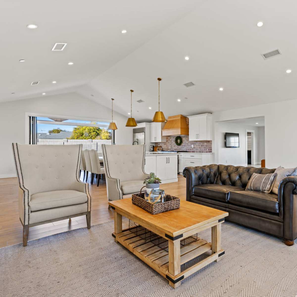 A modern living room with a leather sofa, wooden coffee table, and a dining area featuring upholstered chairs and pendant lights.