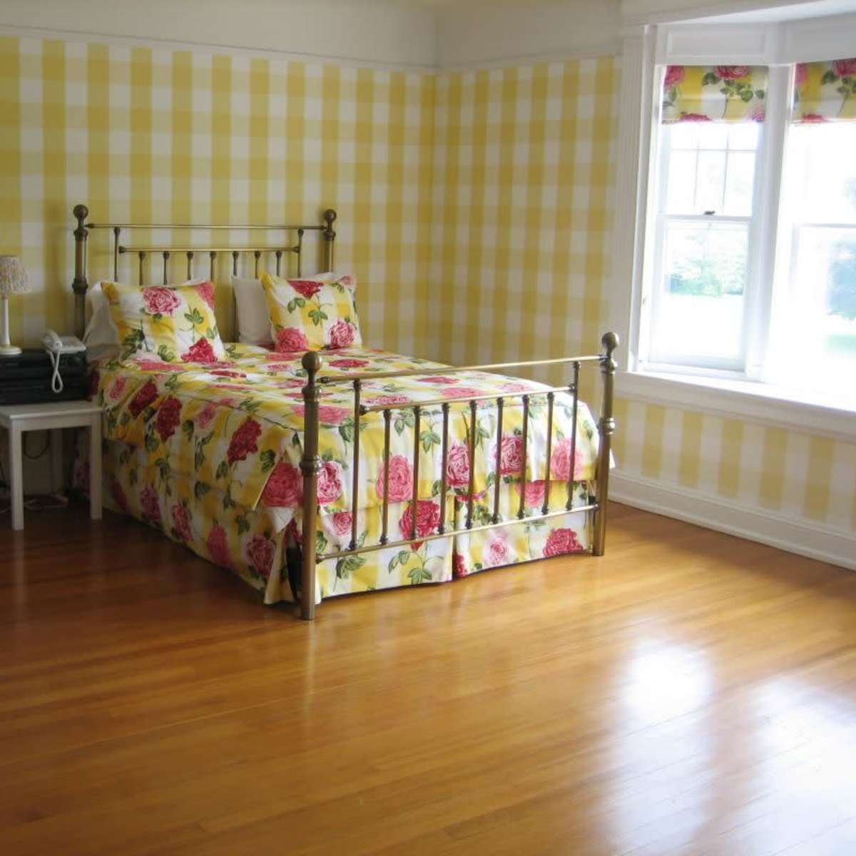A bedroom with a bed covered in a floral pattern quilt, situated on wooden flooring, against a yellow checkered wall.