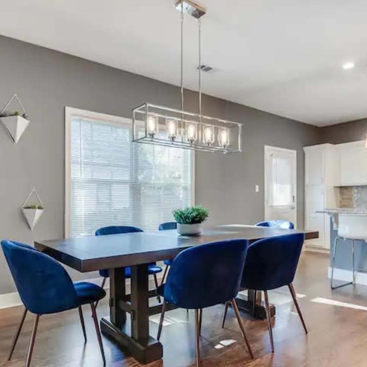 A modern dining area features a large wooden table surrounded by blue chairs, illuminated by a geometric light fixture above, with a kitchen visible in the background.