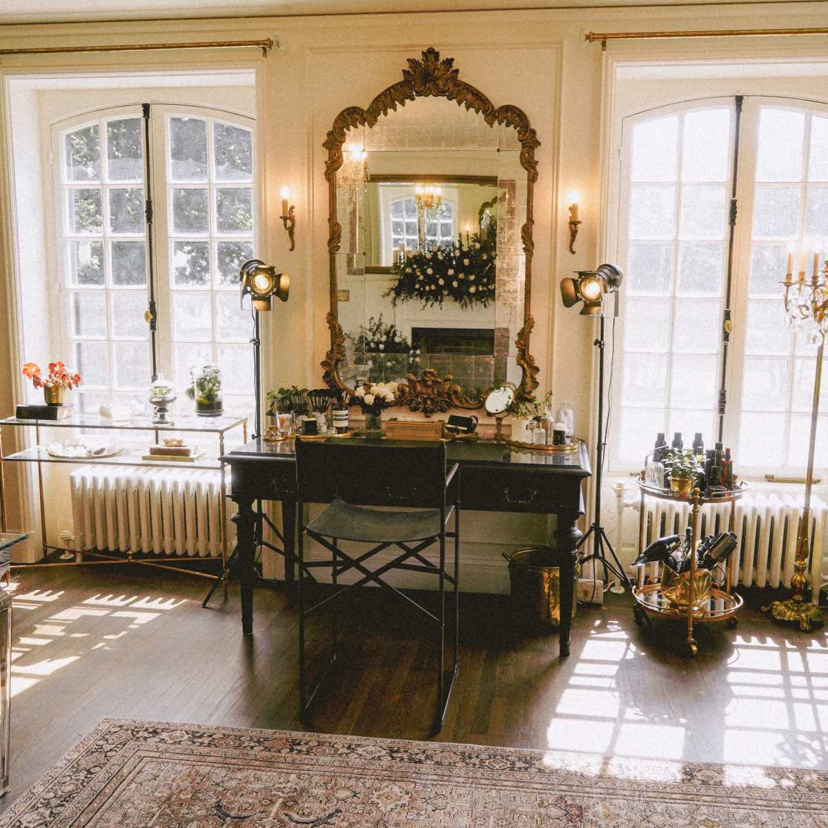 A well-lit room features a vintage vanity table with a large ornate mirror, surrounded by decorative plants and various beauty products.