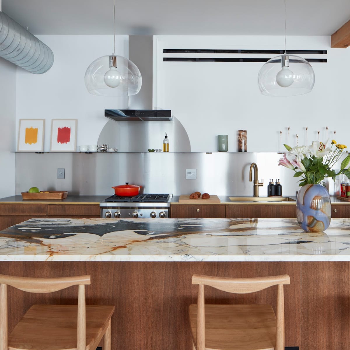 A modern kitchen features a large marble countertop, wooden cabinetry, and hanging light fixtures above a cooking area with a stove and decorative items.