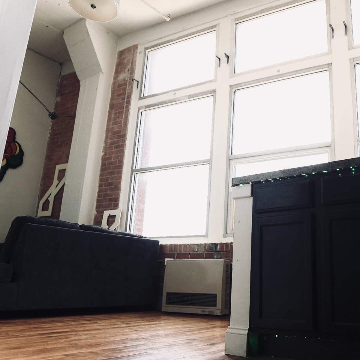 A minimalist indoor space featuring large windows, a dark kitchen countertop, and a gray sofa against a brick wall.