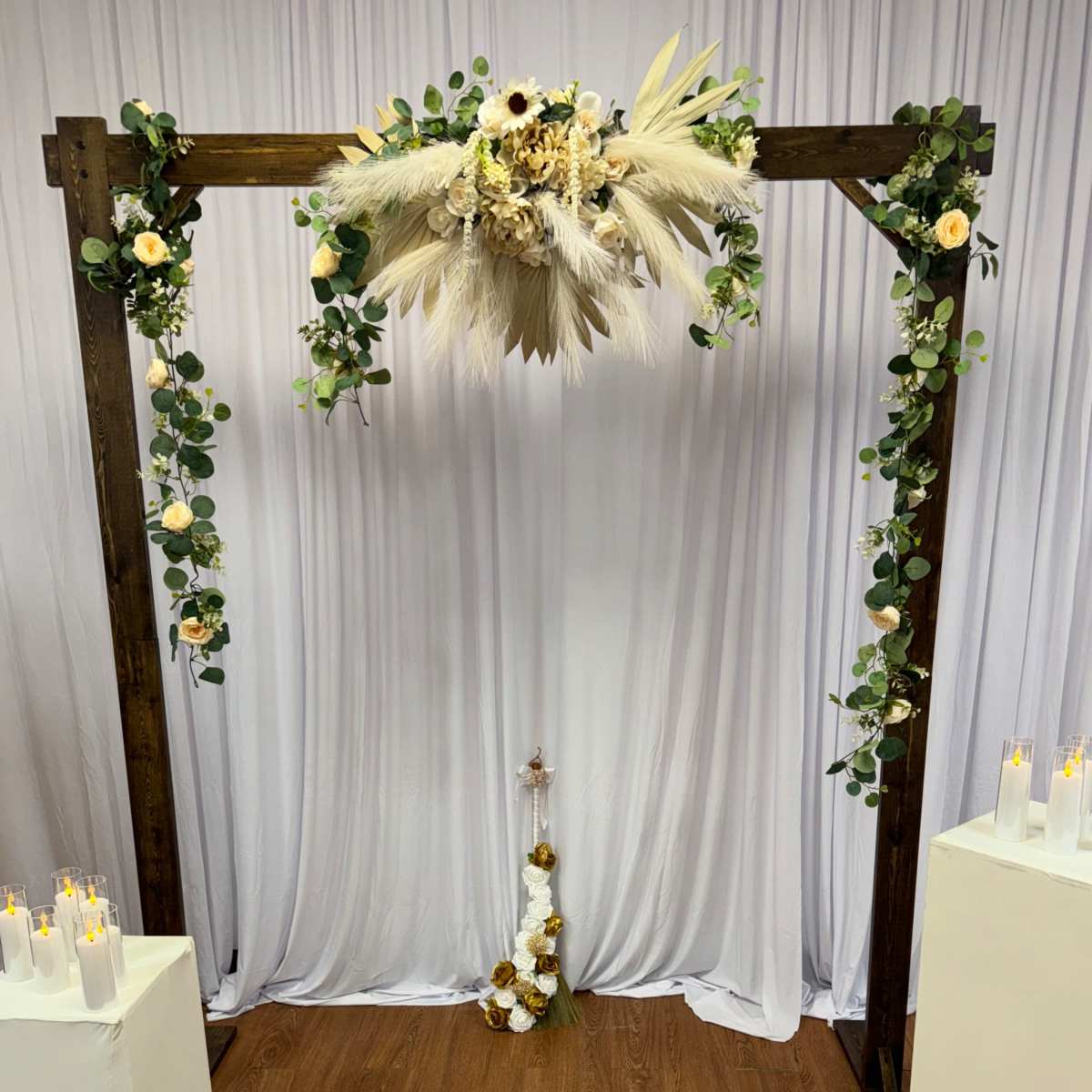 A wooden arch adorned with floral arrangements and greenery is set against a white backdrop, flanked by white pedestals and candles on the floor.