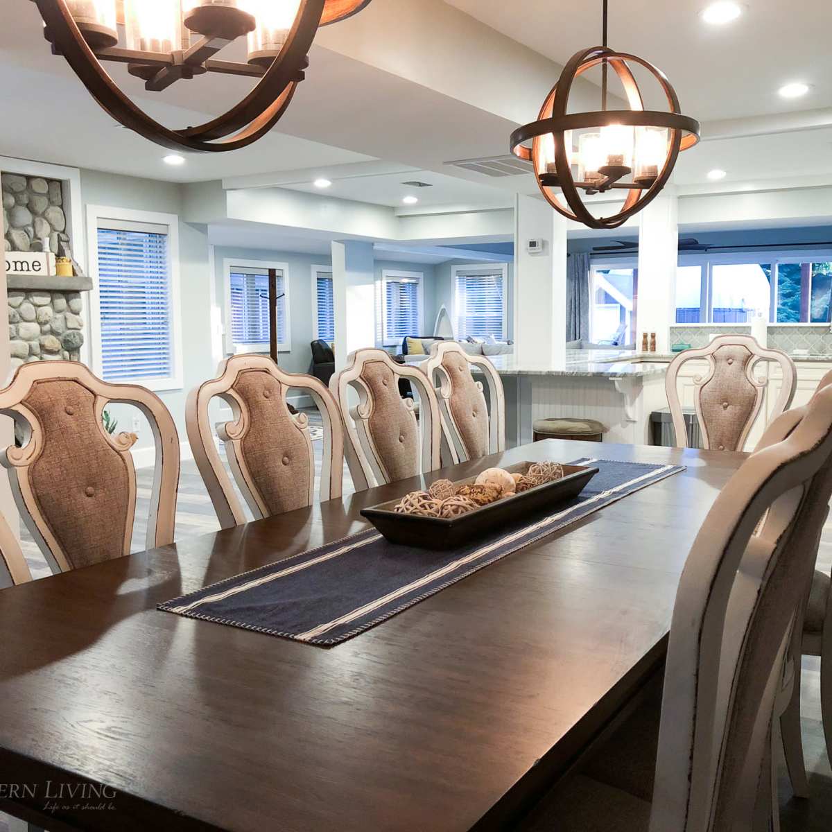 A large dining table with upholstered chairs is set in a well-lit room featuring a modern kitchen in the background.