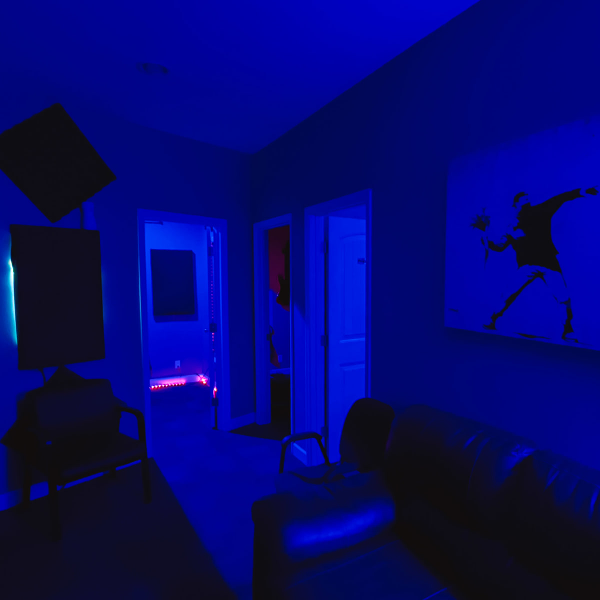 A room illuminated by blue lighting, featuring a black leather sofa, a painting on the wall, and doorways leading to other spaces.