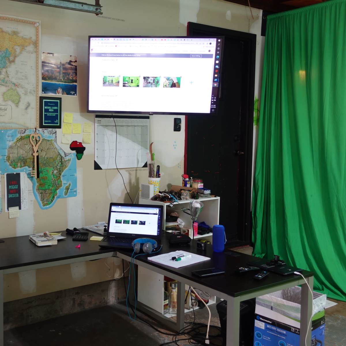 A garage workspace with maps on the walls, a desk with a laptop, a monitor mounted on the wall, and a green backdrop.