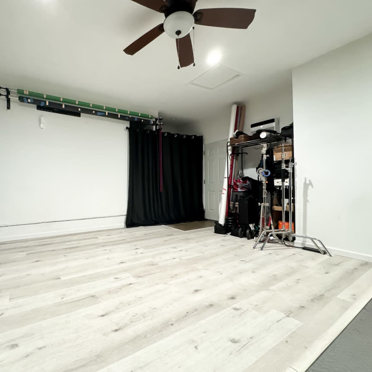 An empty room with light wood flooring, a ceiling fan, black curtains, and equipment neatly arranged against the wall.