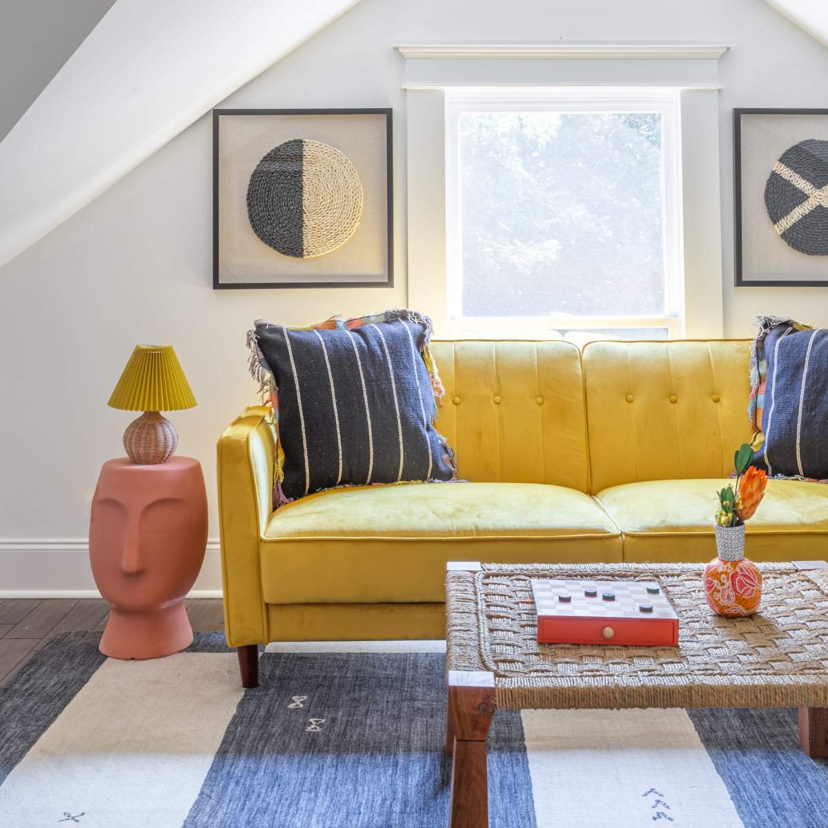 A brightly colored yellow couch with decorative pillows, a small side table shaped like a face, and framed artwork, all in a well-lit attic living space with sloped ceilings and a window.
