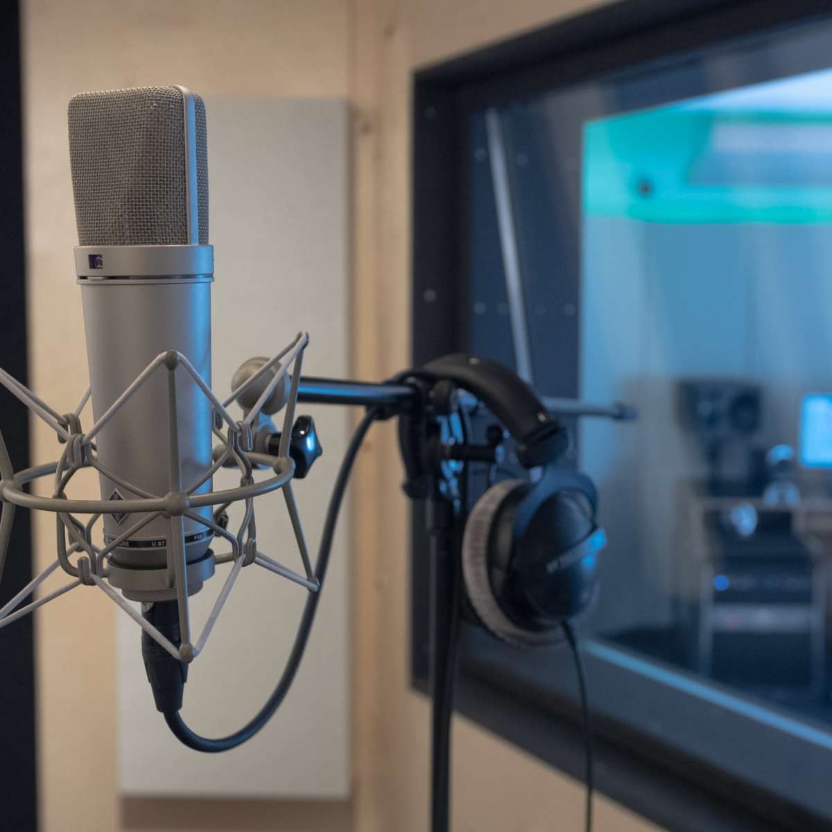 A microphone in the foreground with headphones attached, while a person works at a computer in a sound recording studio in the background.