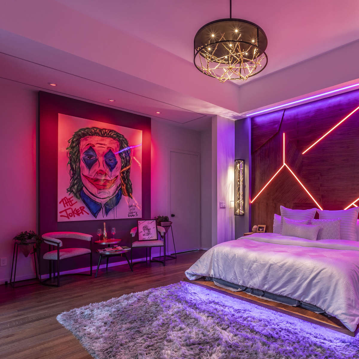A modern bedroom features a large bed with a light blanket, colorful wall art of the Joker, and neon lighting accenting the walls.