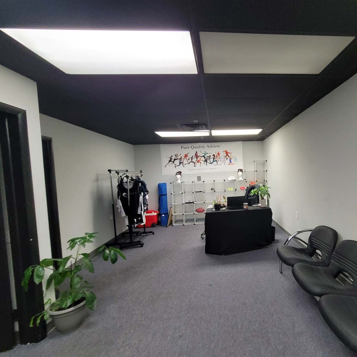 A spacious office area with a black table, seating for three, and shelves displaying various items, along with workout equipment and plants.