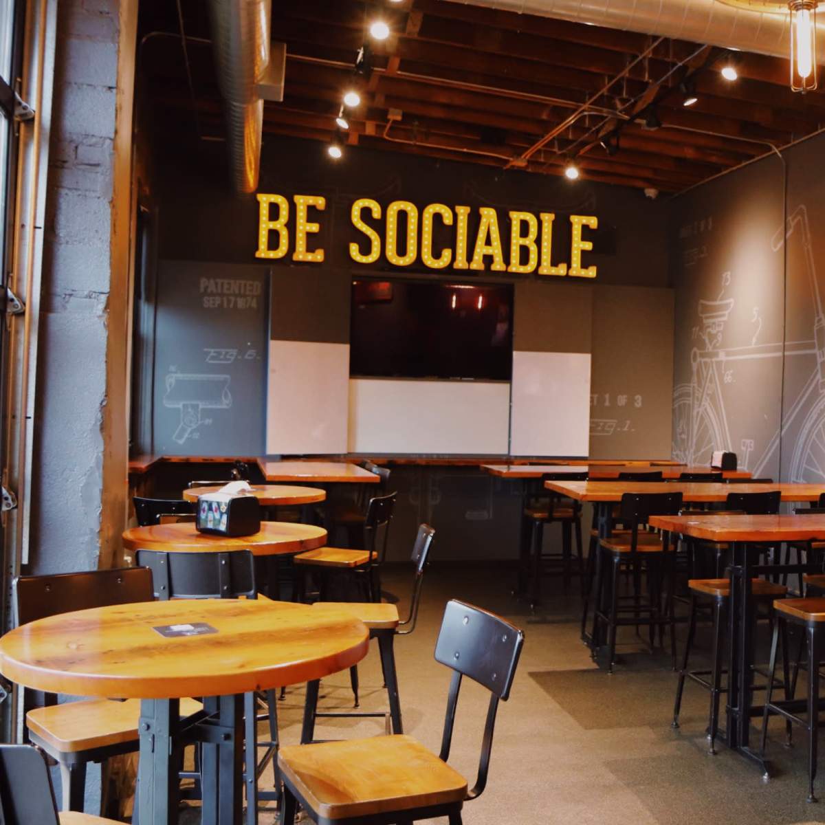 A restaurant interior with wooden tables and chairs, featuring a wall with the phrase "BE SOCIABLE" illuminated above a TV screen and whiteboard.