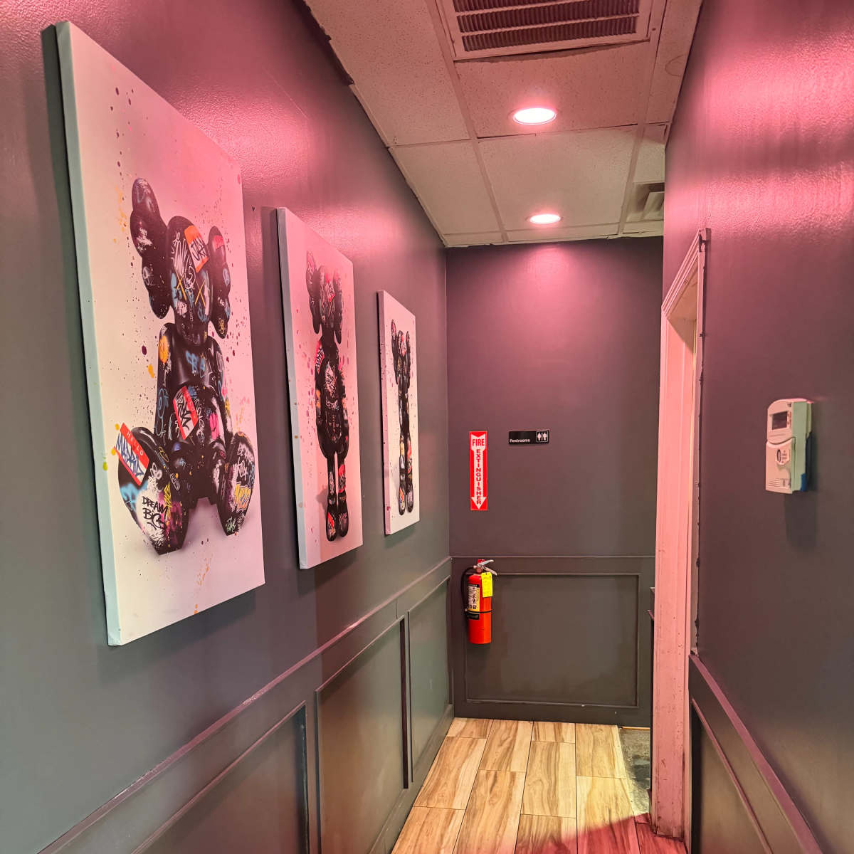 The narrow hallway features three colorful artworks of bear-like figures on the walls, with a fire extinguisher mounted next to a door at the end.
